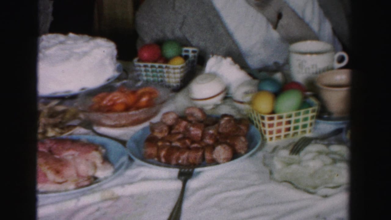 Mid-1950s Easter table in Albany, New York, showing sausages, ham, frosted cake, and scattered multi-colored eggs on a white cloth; partial figure at frame edge