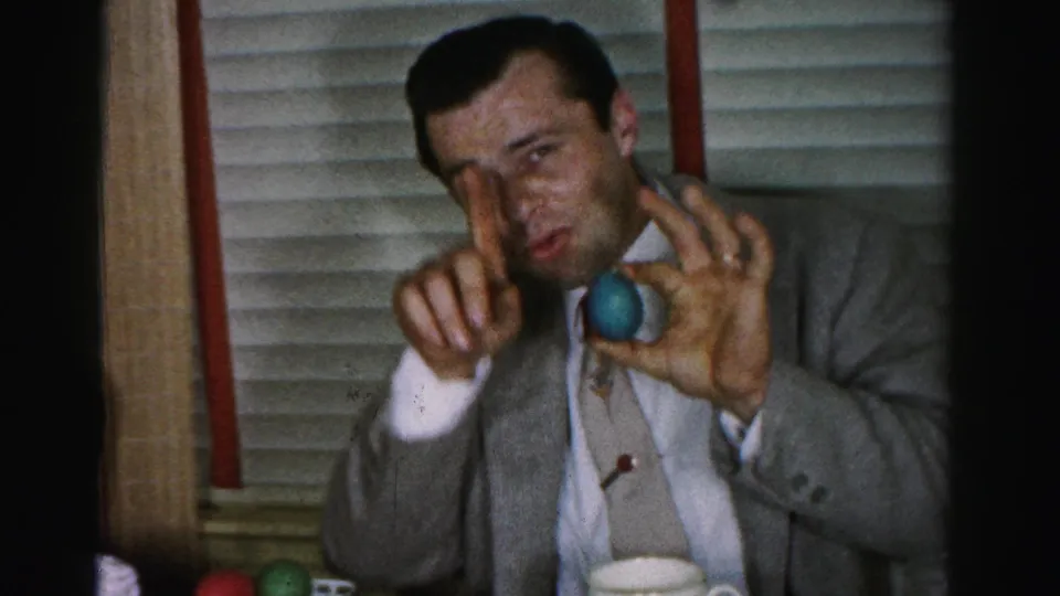 Unidentified man in a mid-1950s grey suit jacket holds a blue Easter egg toward the camera inside what appears to be an Albany, New York home, c. 1955–1956
