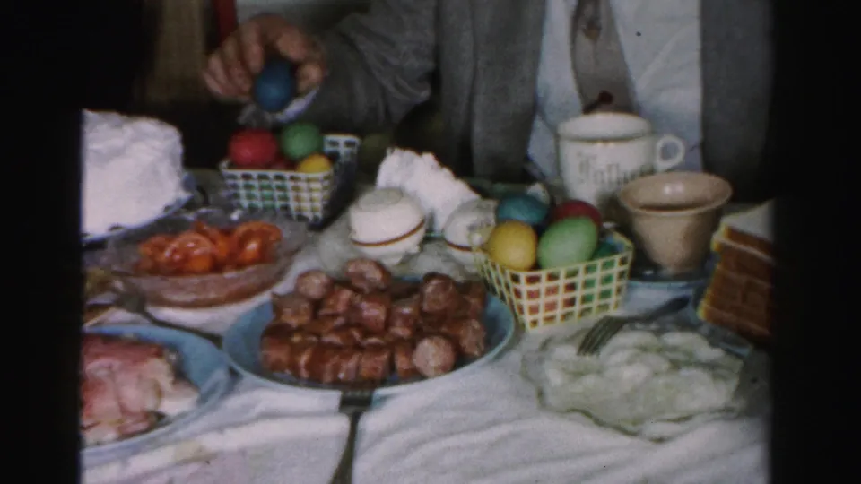 Mid-1950s Easter table in Albany, New York, showing checkered baskets with red, green, and yellow dyed eggs, sausages, ham, and ceramic cups; partial figure interacting with eggs at upper frame