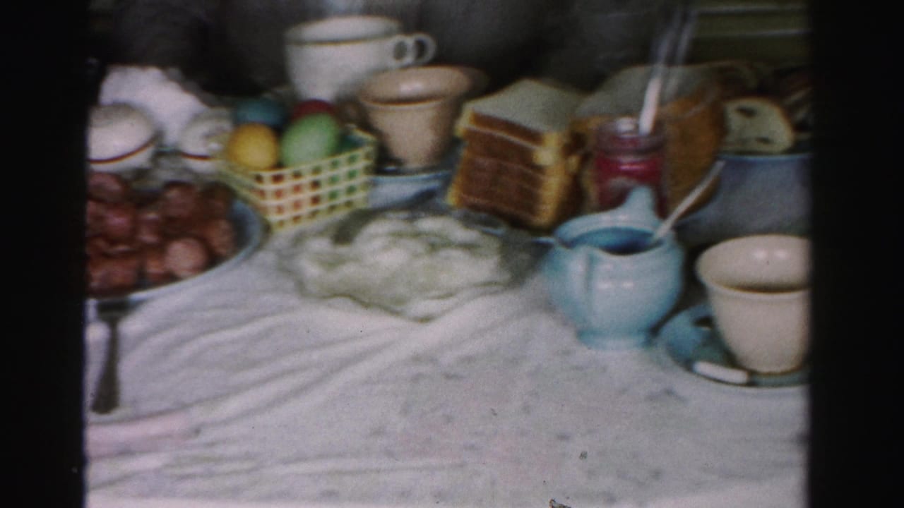Close view of dining table with Easter basket and colored eggs, stacked toast, blue ceramic creamers and white mugs on a white tablecloth.