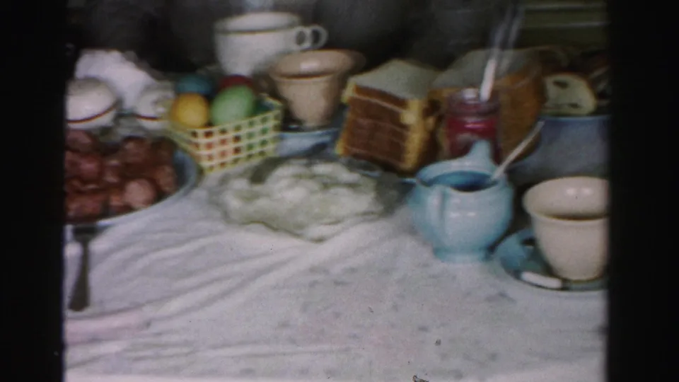 Close-up of 1950s holiday dining table showing Easter basket with colored eggs, stacked toast, blue ceramic creamers, white mugs and dark round food items on white tablecloth.