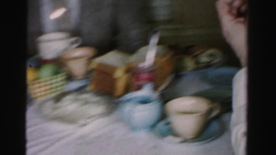 Motion-blurred 8mm pan frame of the Easter table spread — colored egg basket, blue creamer and toast glimpsed through camera movement, c. 1955–1956.