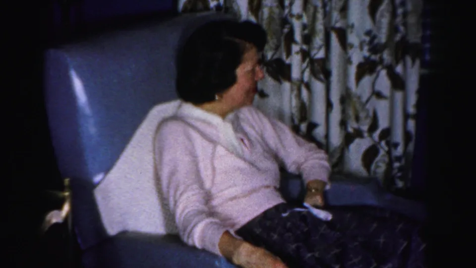 Mid-1950s American domestic interior — woman in pink sweater resting in blue armchair, floral curtain behind; may be an Albany, New York family home, c. 1955–1956.