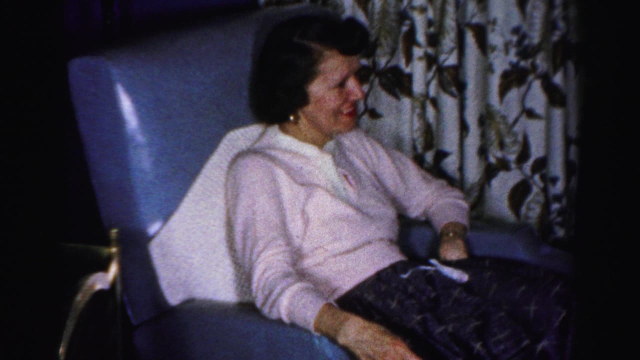Adult woman in a pink sweater sits smiling in a blue armchair in a 1950s Albany living room setting.