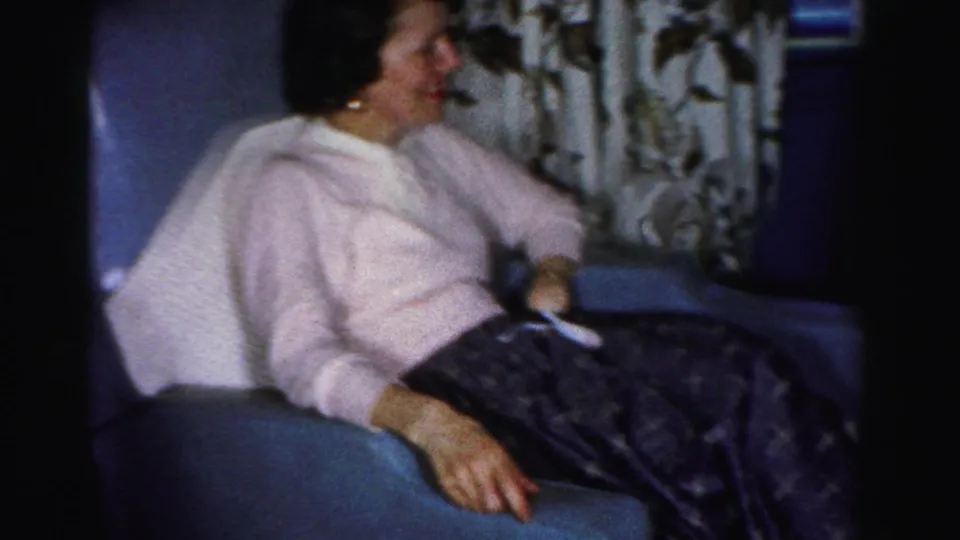 Adult woman in a pink sweater sits in a blue armchair, turned toward activity in the Albany home interior.