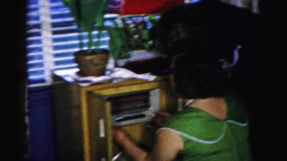 Adult woman in a green dress adjusts a wooden console radio in a 1950s Albany living room with blinds and a plant.
