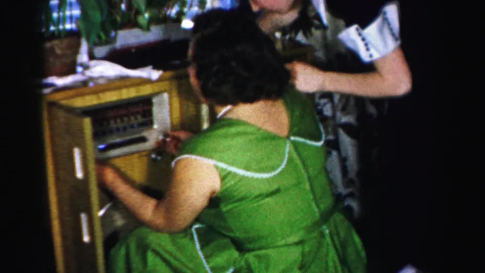 Adult woman in a green dress works the console radio while another adult stands nearby in the living room.