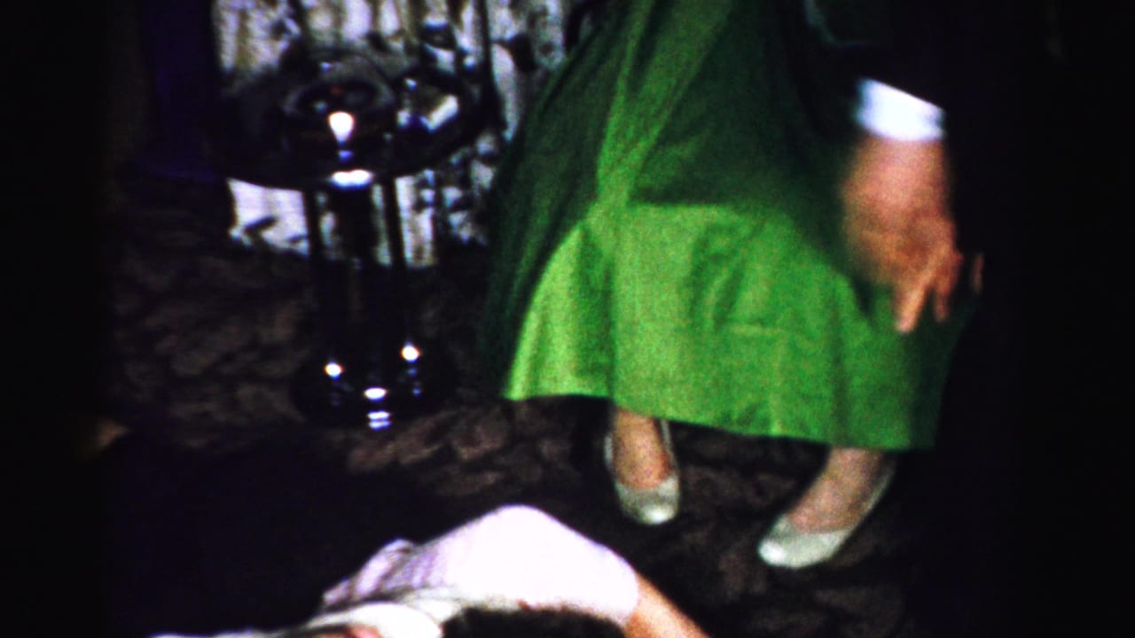 Low-angle view of a woman in a green dress standing over a person in white clothing on a dark patterned carpet, interior setting.