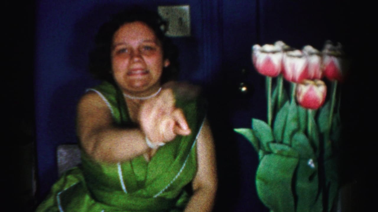 Woman in green sleeveless dress gesturing beside a large bouquet of pink and white tulips against a dark blue background.