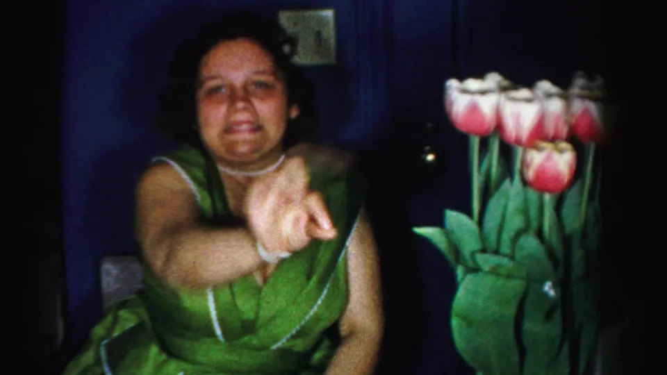 Mid-1950s woman in a green sleeveless dress gestures toward a bunch of pink and white tulips in a domestic interior, 8mm home movie, Albany, New York, c.1956.
