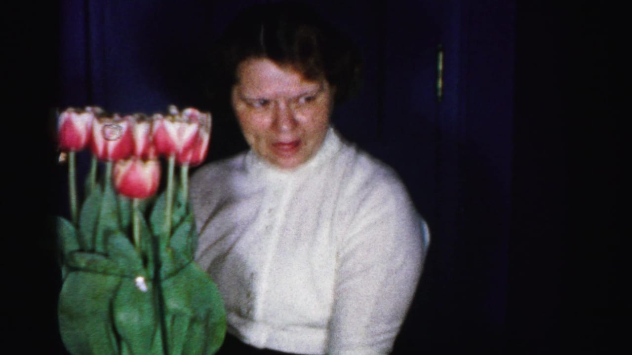 Woman in white knit blouse with pearl necklace standing beside a large bouquet of pink and white tulips against a dark blue background.