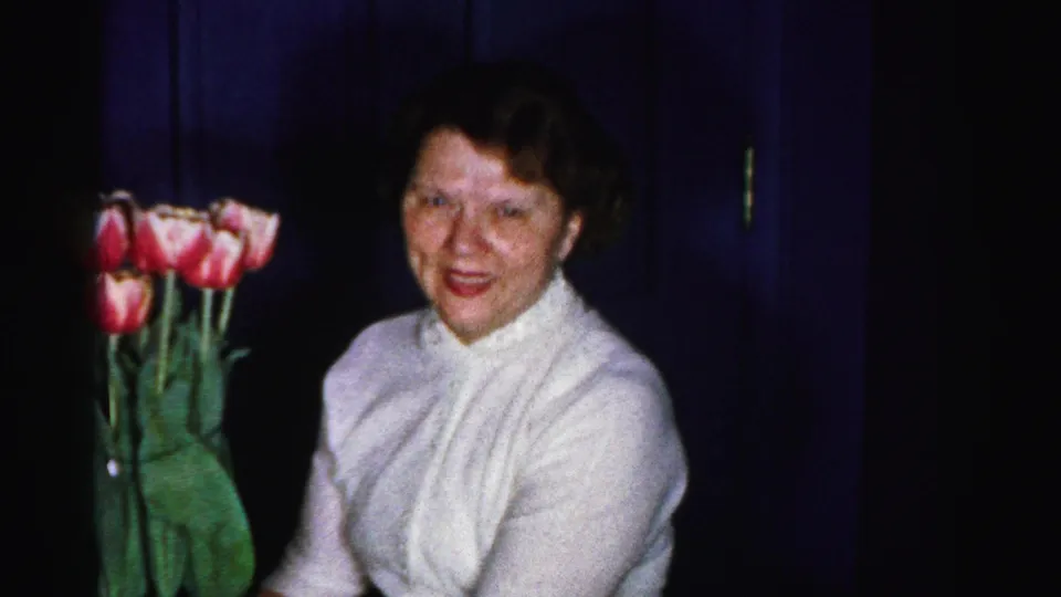 Mid-1950s woman in a white knit blouse smiles toward the camera beside pink and white tulips in a domestic interior, 8mm home movie, Albany, New York, c.1956.