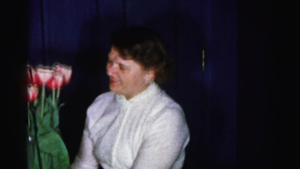 Woman with short reddish-brown hair in a white textured blouse holds tall-stemmed pink and red tulips; dark blue interior behind her; 8mm home movie, Albany, New York area, mid-1950s.
