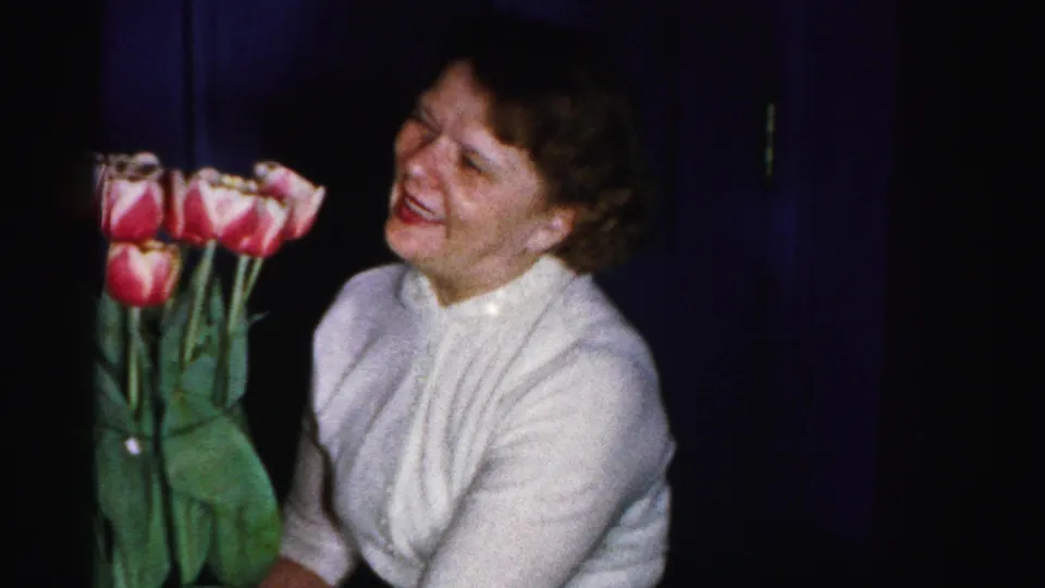 Candid 8mm home movie frame of a woman in a white blouse laughing beside pink and red tulips; unguarded expression consistent with intimate family filming; Albany, New York area, mid-1950s.