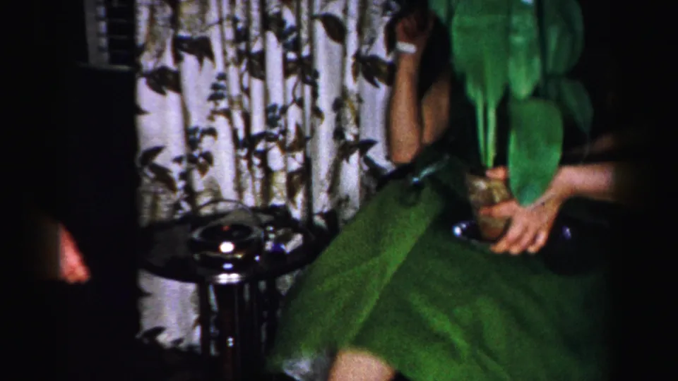 Woman in a green skirt handling a vase beside botanical-print curtains and a dark lamp base in what appears to be a 1950s American home.
