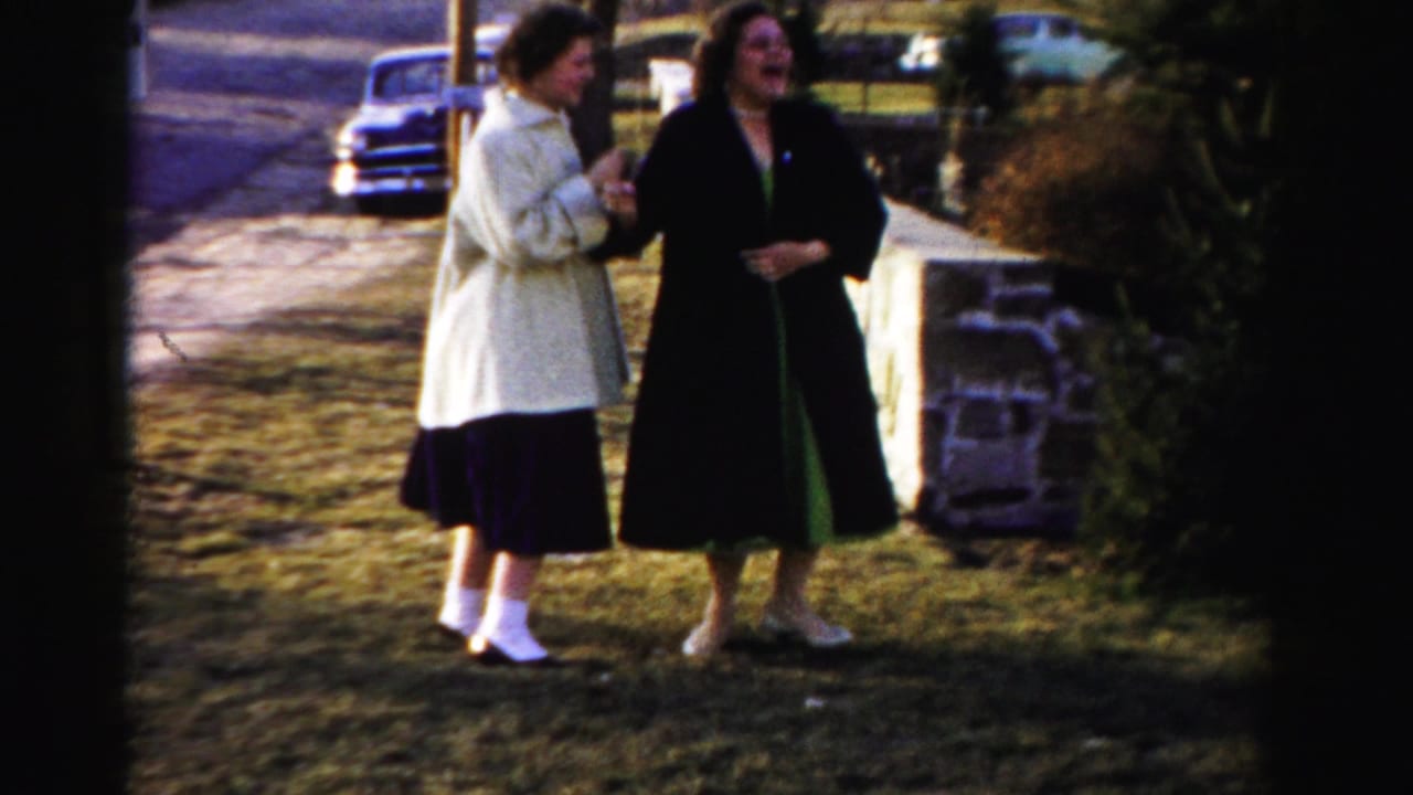 Two women in 1950s-era coats, one cream and one dark, laughing together on a dormant lawn with a vintage automobile and low stone wall in background, Albany, New York area, circa 1955-1956.