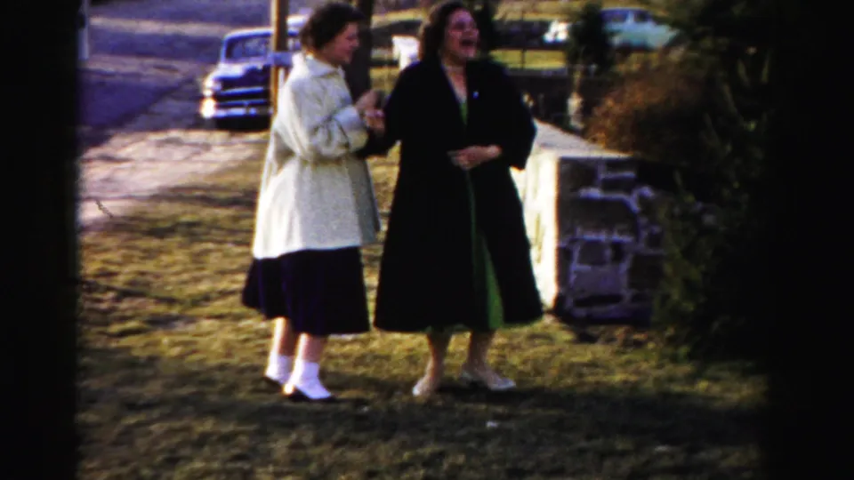 Two women in 1950s-era coats, one cream and one dark, laughing together on a dormant lawn with a vintage automobile and low stone wall in background, Albany, New York area, circa 1955-1956.