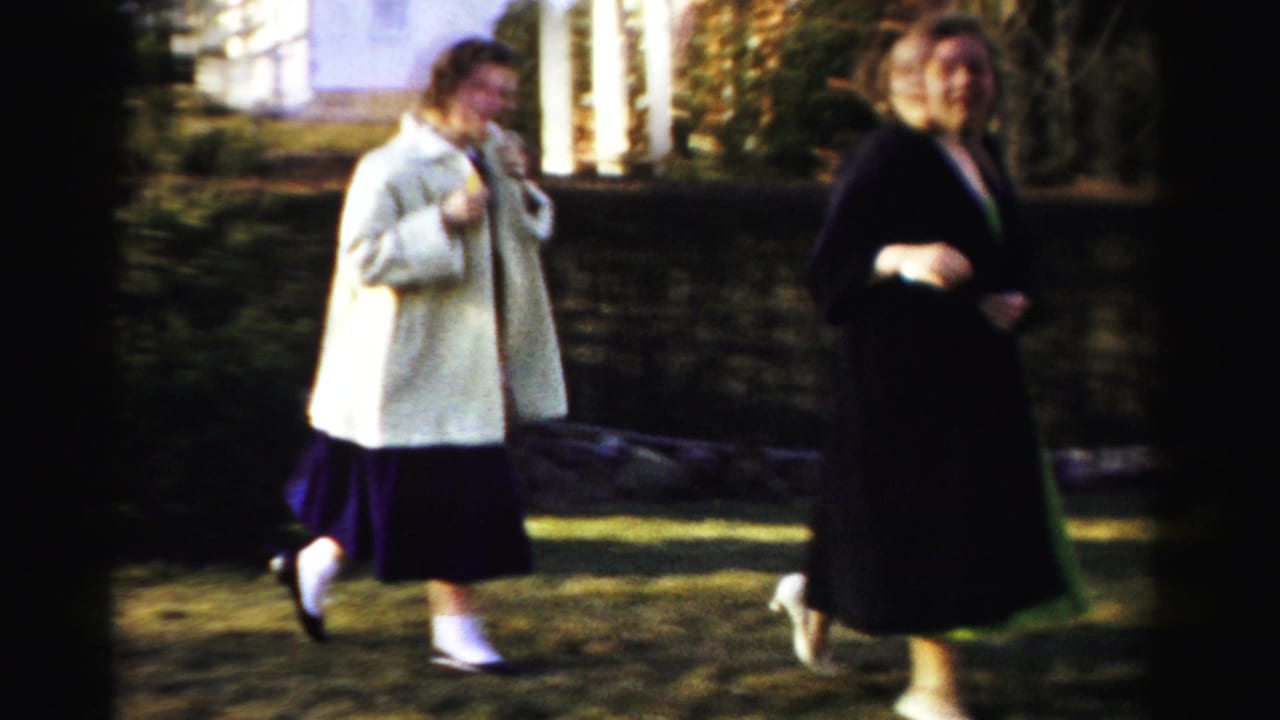 Two women in 1950s-era structured coats, one cream and one dark, walking or running across a dormant lawn with a white building and evergreen trees in background, circa 1955-1956.