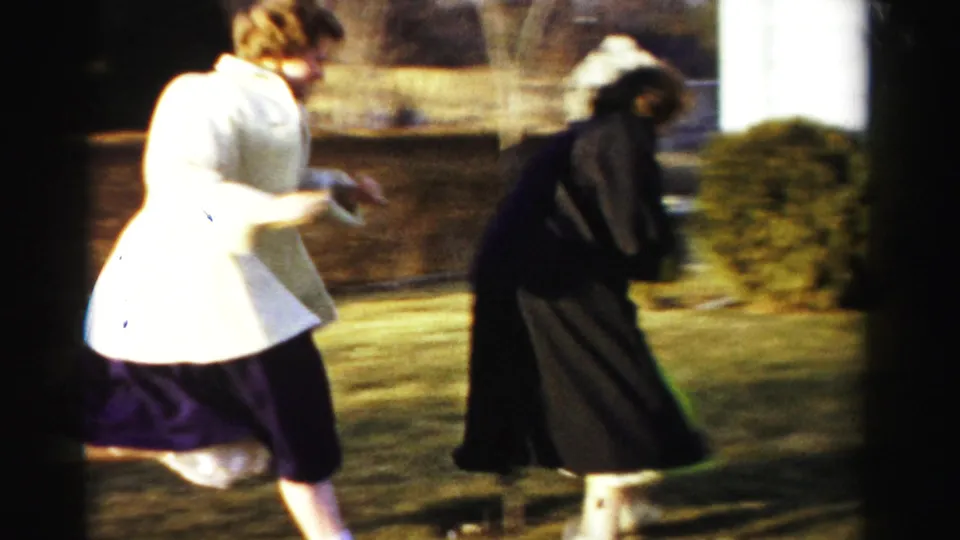 Two women in 1950s-era coats caught mid-motion on a sparse lawn, one bending forward with motion blur visible, evergreen shrubs and a white structure in background, circa 1955-1956.