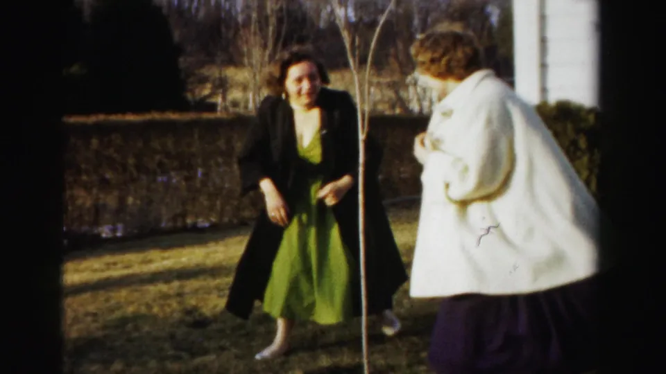 Two women pause on a dormant lawn, one in a green dress and one in a cream coat, with a low stone wall, bare sapling, and white structure in background, circa 1955-1956, Albany, New York area.