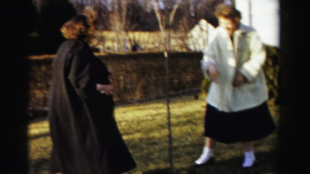 Two women in coats stand close together in a suburban backyard; white building visible at upper right.