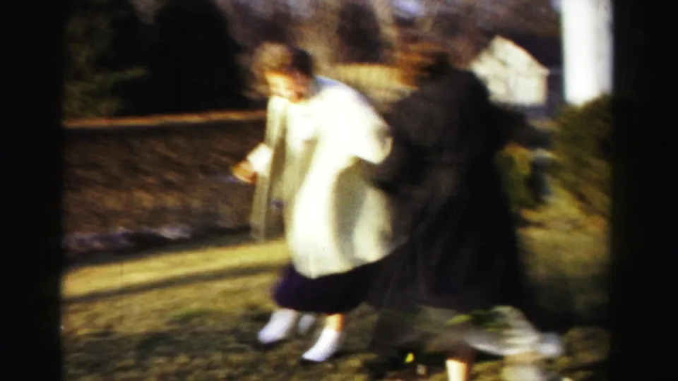 Motion-blurred 8mm frame of two young women in coats engaged in playful mock-boxing; circa 1955, suburban backyard setting.