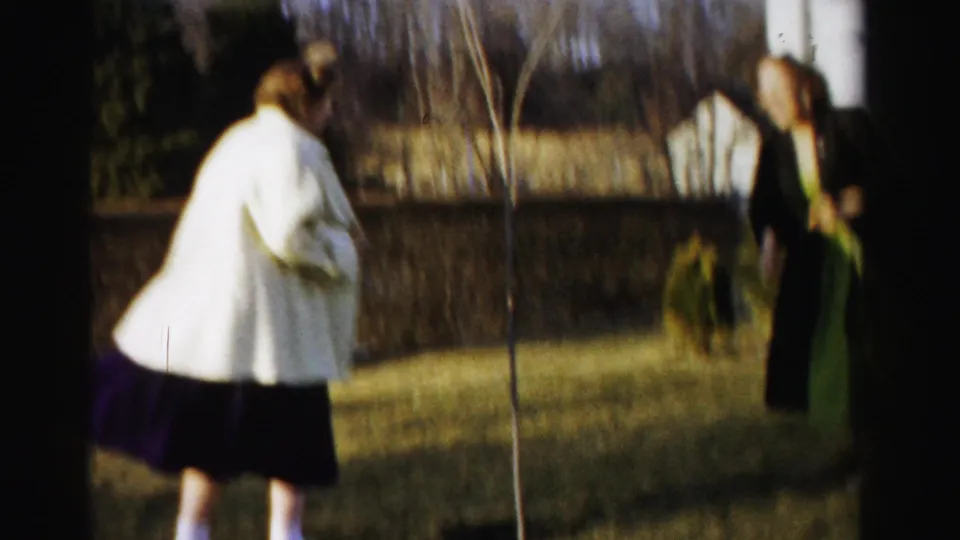 Two young women in 1950s coats stand separated on a dormant suburban lawn with a thin young sapling between them, Albany, New York, circa 1955.