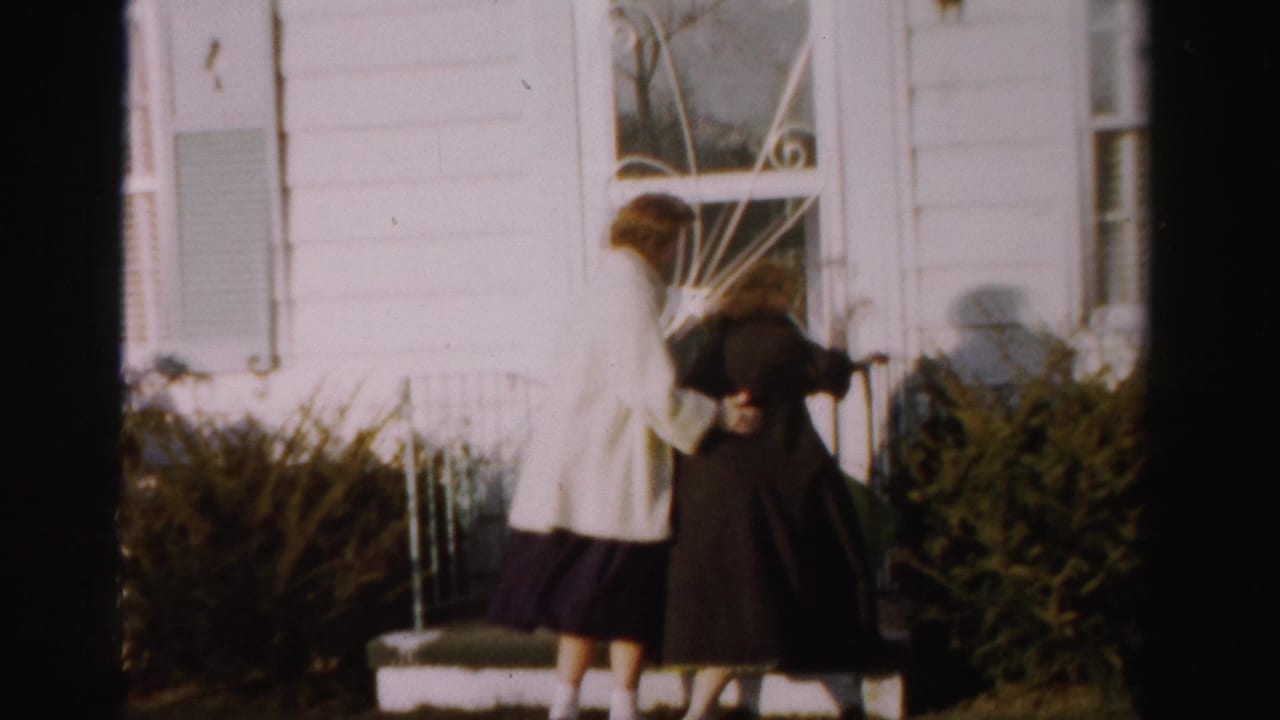 Two women in 1950s coats, one light and one dark, climbing porch steps of a white clapboard house with ornate ironwork railing, backs to camera.