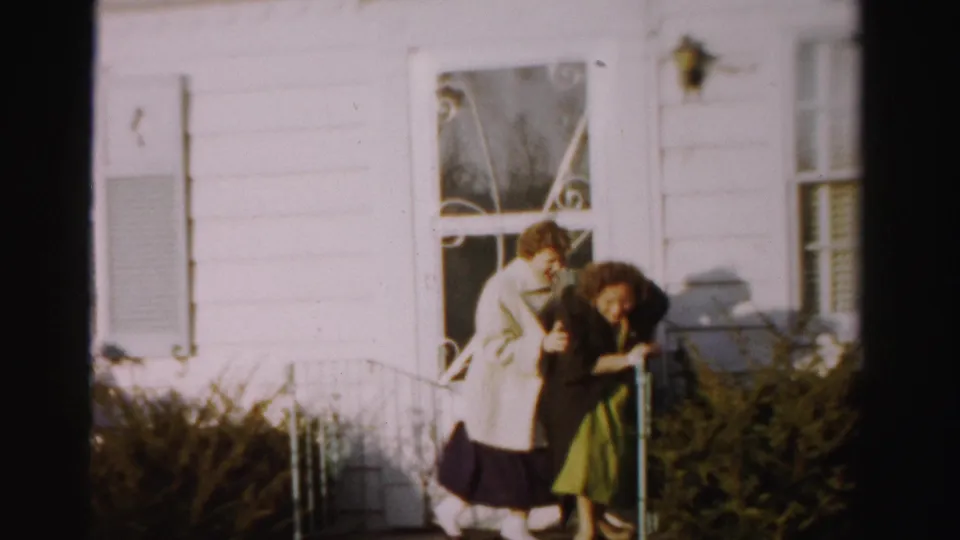Two women laughing and steadying each other near a porch doorway in 1950s outerwear; ornate ironwork railing and wall-mounted lamp visible, circa mid-1950s.