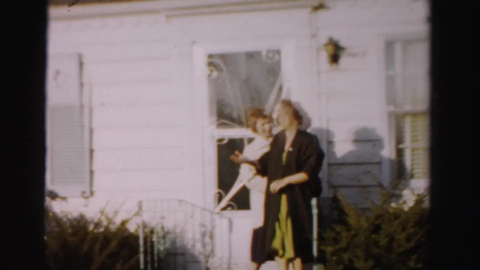 Two adult women in 1950s outerwear laughing outside a white clapboard house with decorative ironwork railing and shuttered windows; faces clearly visible, possibly Albany, NY area.