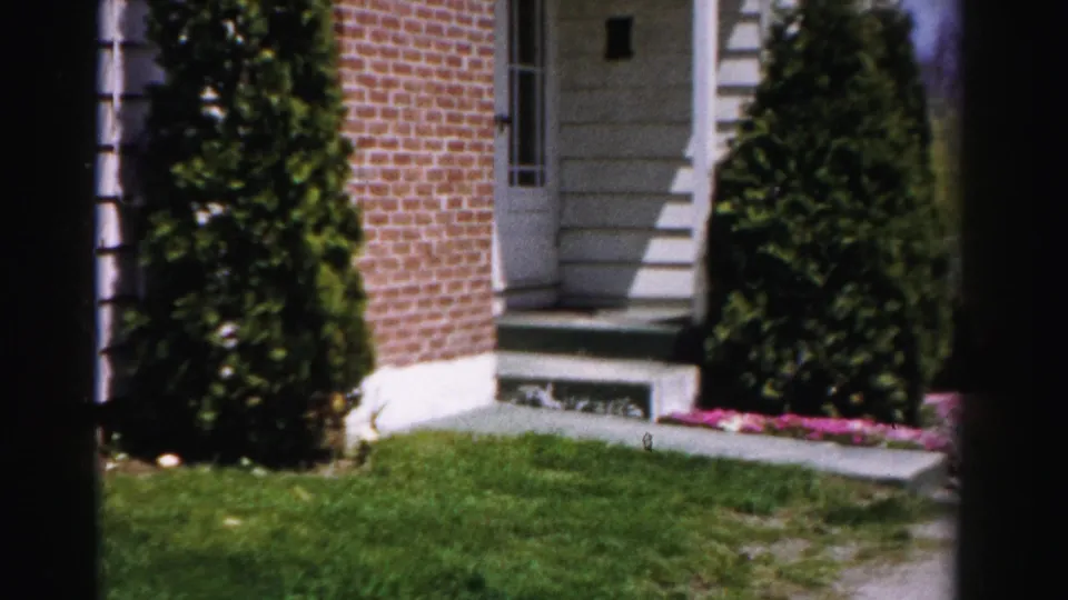 Front entry steps and door of a brick residential house with white pilasters and flanking arborvitae-style shrubs, from a mid-1950s 8mm home movie, Albany, New York.