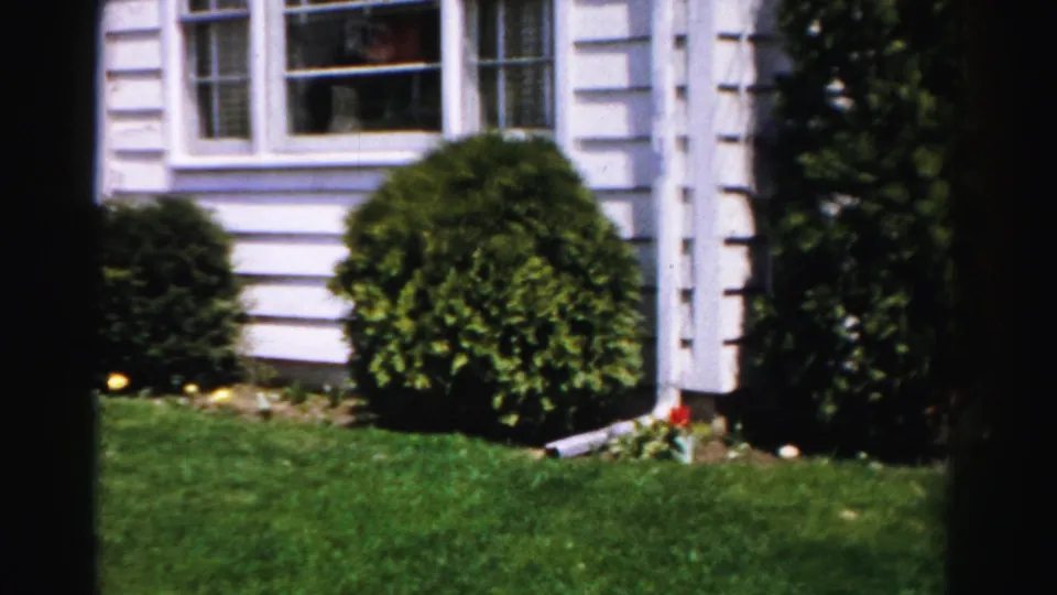 8mm color frame, c.1955–56, showing white clapboard siding, white-framed windows, a manicured globe shrub, and a bright summer lawn at a residential property in Albany, New York.
