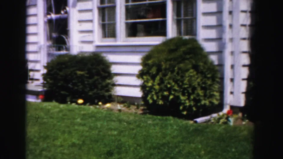 Closing 8mm frame, c.1955–56, showing the suburban home's side elevation with a manicured globe shrub, bright summer lawn, and flowering border, Albany, New York area.