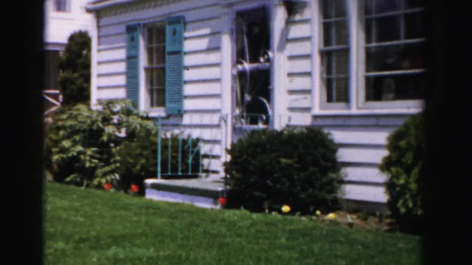 8mm home movie pan showing white clapboard house facade with teal shutters, front entry steps with railing, foundation shrubs and orange-yellow flowers, Albany NY area, mid-1950s.