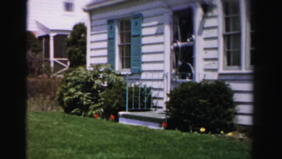 Final pan frame of 8mm home movie showing white clapboard house with teal shutters, front steps with teal railing, foundation shrubs and orange-yellow blooms, Albany NY area, mid-1950s.
