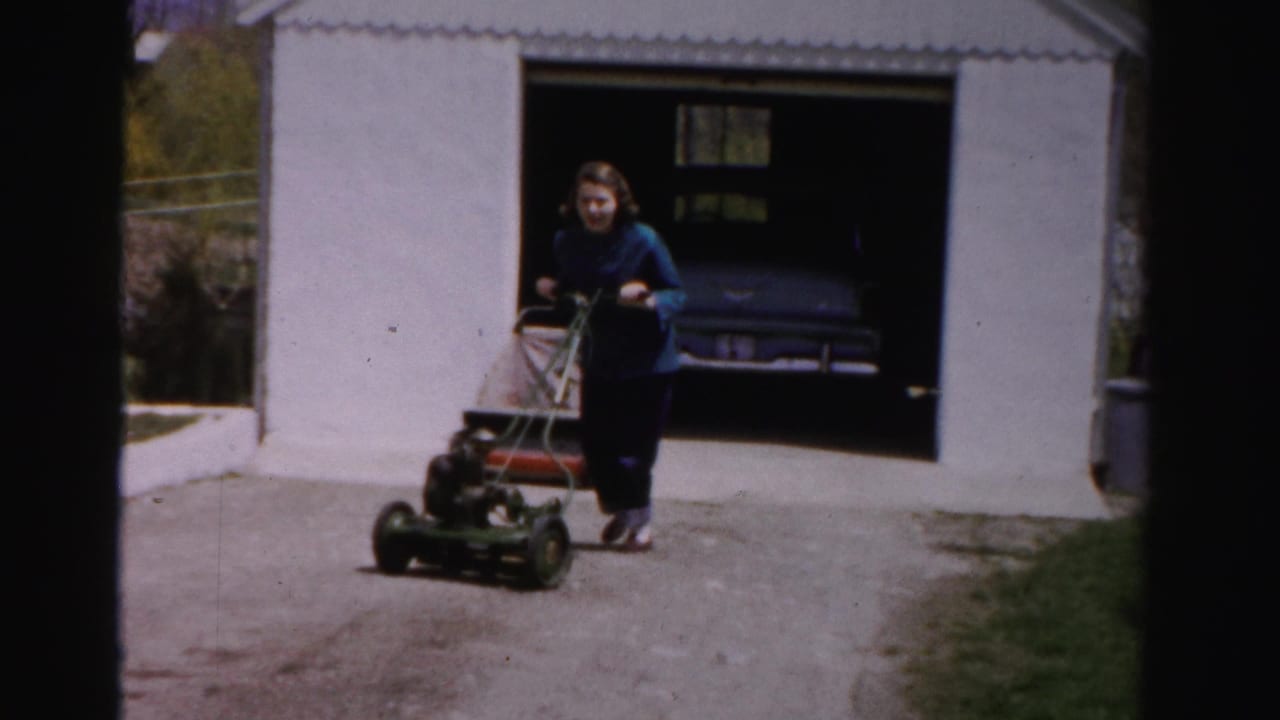 Mid-1950s woman in blue sweater and dark slacks operating a green rotary power mower near a white suburban garage, Albany, New York area, circa 1955–1956.