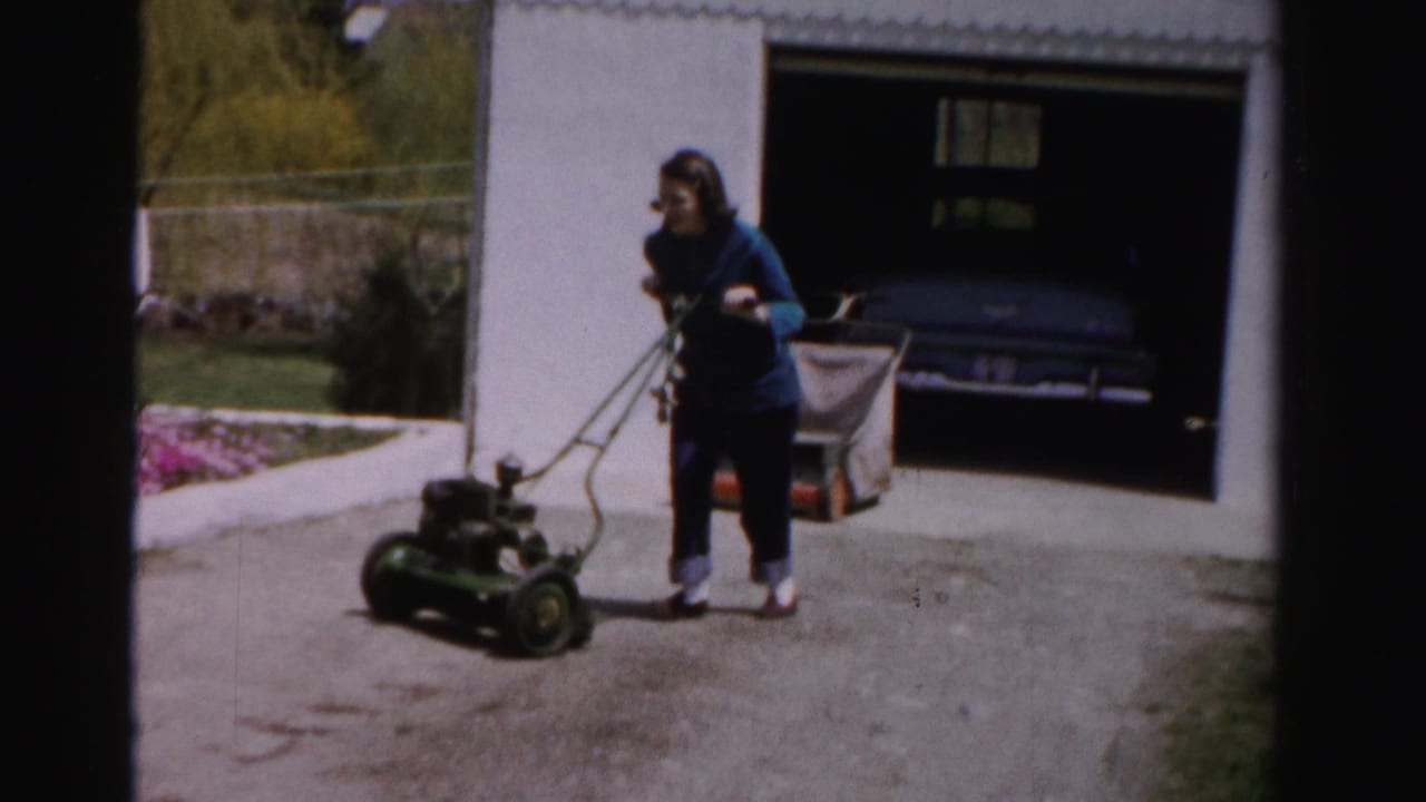 Woman in blue top guides green rotary mower with rear grass catcher on driveway; white garage with car inside visible at left, flowering spring trees at right.