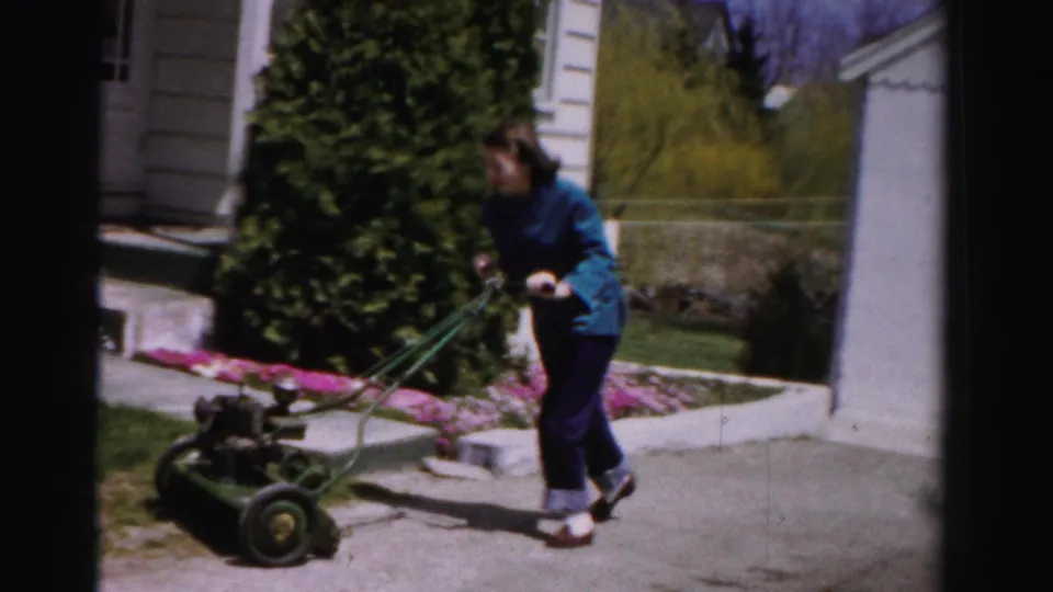 Mid-1950s woman mowing near a suburban garden bed of pink-blooming shrubs; mature deciduous trees and white clapboard house in background, Albany, New York area, circa 1955–1956.