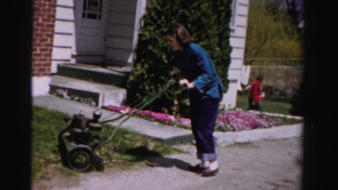 Woman in blue jacket pushes green gas lawnmower on suburban lawn; small red-clothed figure visible in background near house steps.