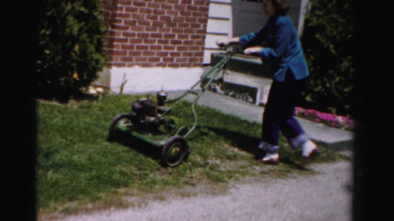 Woman in blue jacket pushes vintage gas push mower beside brick-and-white-trim suburban house; Albany, New York area, circa mid-1950s 8mm home movie.