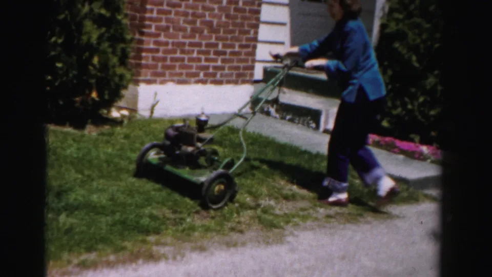 Woman in blue jacket pushes vintage gas push mower beside brick-and-white-trim suburban house; Albany, New York area, circa mid-1950s 8mm home movie.