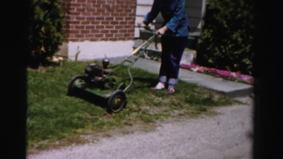 Mid-shot of woman in blue jacket operating vintage gas push mower along garden border; brick suburban house exterior, Albany, New York area, circa mid-1950s.