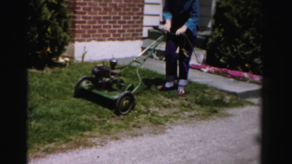 Woman in blue jacket with vintage gas push mower on lawn beside brick house and flower bed; Albany, New York area, circa mid-1950s 8mm home movie.