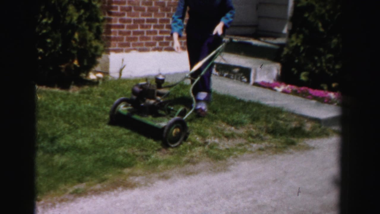 Woman in blue top and dark pants leaning over a vintage green push mower near a brick house with flower bed, suburban Albany, New York area, mid-1950s 8mm footage.