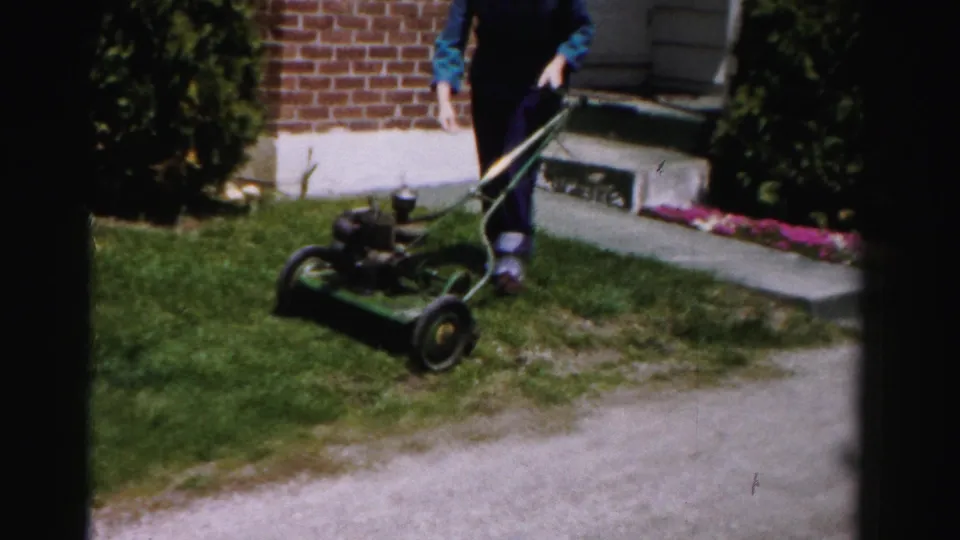 Woman in blue top and dark pants leaning over a vintage green push mower near a brick house with flower bed, suburban Albany, New York area, mid-1950s 8mm footage.
