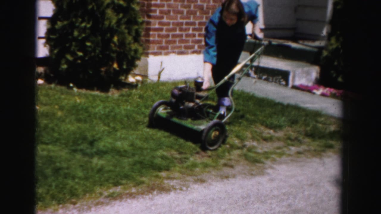 Woman in blue bent over a green push mower near a brick wall and trimmed evergreen shrub on a sunny lawn.