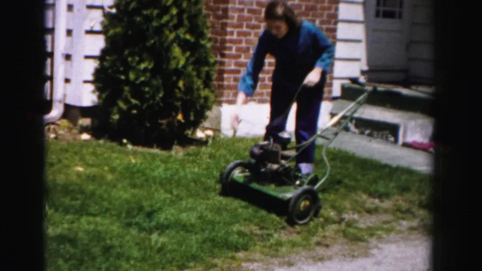 Woman in blue top and dark pants bending over a vintage green push mower beside a brick house with white door trim and trimmed shrubs, mid-1950s 8mm home movie.