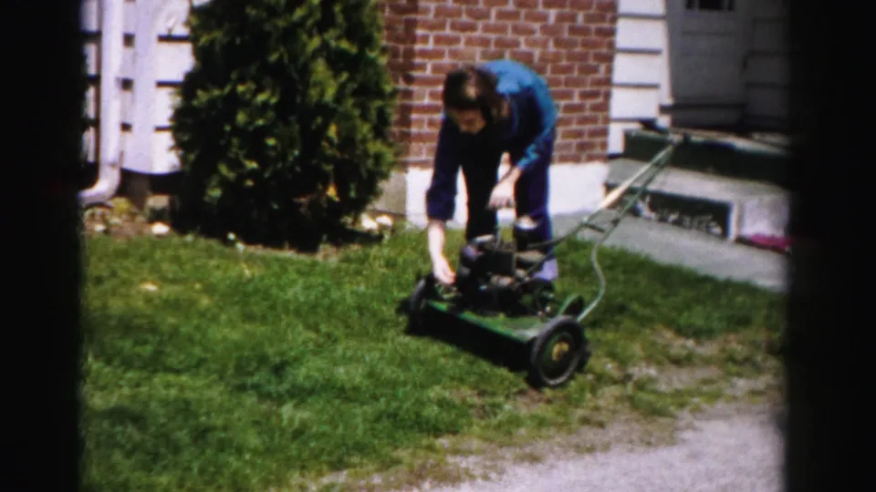 Woman in blue top bent over a vintage green rotary push mower near a brick house, white fence or lattice structure at left, mid-1950s 8mm suburban home movie.