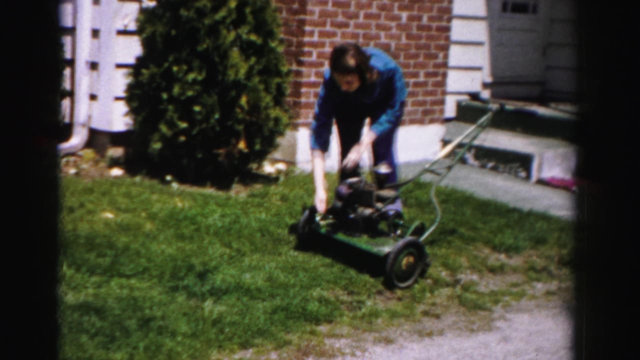 Woman in blue jacket leaning forward to push a green rotary mower across a front lawn, brick house and evergreen shrub behind her.