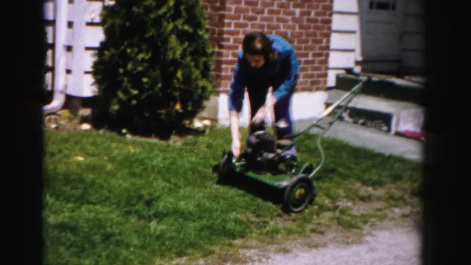 Woman in blue jacket pushes a green gas-powered rotary mower across a suburban front lawn; brick house with white trim visible; Albany, New York, mid-1950s.